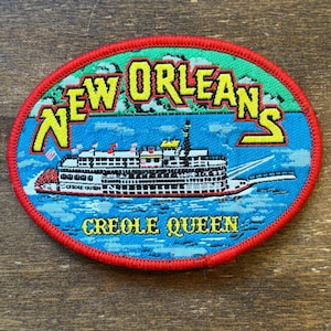 Peut inclure: Écusson ovale avec une bordure rouge. Il présente les mots "NEW ORLEANS" en lettres jaunes. Il représente un bateau à vapeur sur de l'eau bleue, avec les mots "CREOLE QUEEN" en dessous. Le fond est vert et bleu.