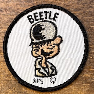 May include: A circular embroidered patch with a white background and a black stitched border. It features a cartoon character wearing a hat with the word BEETLE embroidered in black above the head. The character has tan skin and black outlines.