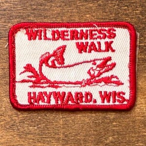 May include: A rectangular embroidered patch with a white background and a red border. The patch features a red stitched graphic of a fish jumping out of water, with the text WILDERNESS WALK above and HAYWARD, WIS. below in red embroidery.