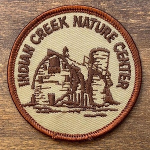 Może przedstawiać: Okrągła naszywka z napisem "INDIAN CREEK NATURE CENTER" w brązowym, łukowatym kroju pisma. Środek przedstawia brązową ilustrację budynku i silosu. Naszywka ma beżowe tło i brązową, szytą obwódkę.