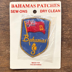 Puede incluir: Un parche azul y dorado de Bahamas con un diseño de bandera roja y caballito de mar. El parche está en una funda de plástico transparente y sujeto a un respaldo de cartón blanco. El texto dice "BAHAMAS PATCHES" y "MADE FOR THE BAHAMAS."