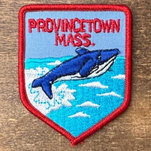 May include: A shield-shaped embroidered patch with a red border. The design features a blue whale leaping out of turquoise water against a light blue sky. The text PROVINCETOWN MASS. is stitched in red letters at the top of the patch.