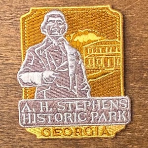 Puede incluir: Parche bordado con borde dorado, que representa a un hombre con traje, un edificio y el texto "A. H. STEPHENS HISTORIC PARK GEORGIA." El fondo es de color beige.