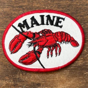 May include: Embroidered patch featuring a red lobster on a white oval background. The word "MAINE" is arched above the lobster in black letters. The patch has a red stitched border and is likely for clothing or accessories.