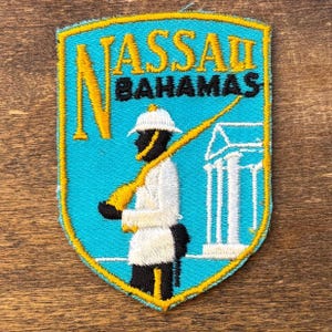 Puede incluir: Parche bordado con forma de escudo, con el texto "NASSAU BAHAMAS" en amarillo y negro. El parche tiene un fondo turquesa y un borde amarillo. Una figura con uniforme blanco y piel negra sostiene un rifle.