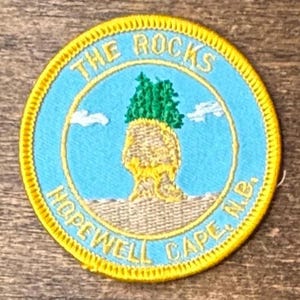 Puede incluir: Un parche bordado circular con un borde amarillo brillante. El centro presenta un cielo azul claro con nubes blancas, una formación rocosa de color tostado y árboles verdes encima. El texto dice THE ROCKS arriba y HOPEWELL CAPE, N.B. abajo.