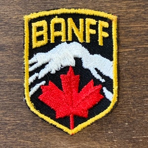 May include: Embroidered patch featuring the word "BANFF" in yellow letters above a white mountain range and a red maple leaf. The patch has a black background with a yellow border. A souvenir item.
