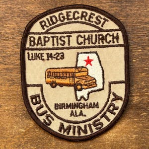 以下が含まれることがあります： 「RIDGECREST BAPTIST CHURCH」と「BUS MINISTRY」の文字が刺繍されたパッチ。パッチにはスクールバスのグラフィックと、赤い星が付いたアラバマ州の輪郭が描かれています。「LUKE 14:23」と「BIRMINGHAM ALA.」の文字も含まれています。