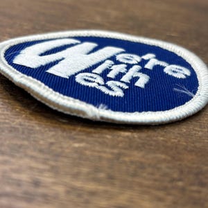 Puede incluir: Un parche bordado circular con un fondo azul profundo y un borde cosido en blanco. El parche presenta el texto bordado en blanco "CHere ith es" dispuesto en un dise&ntilde;o estilizado y apilado sobre el centro de la superficie de tela azul.
