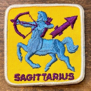 May include: A square embroidered patch with a yellow background and a white border. It features a light blue centaur figure holding a purple bow and arrow. The word SAGITTARIUS is embroidered in purple capital letters at the bottom of the patch.