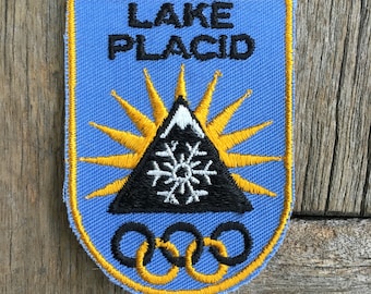 Puede incluir: Un parche bordado en forma de escudo con fondo azul claro y borde amarillo. Presenta el texto negro LAKE PLACID sobre una montaña negra con pico blanco, un copo de nieve blanco, un sol amarillo y tres anillos olímpicos negros.