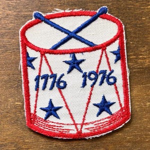 May include: Embroidered patch with a drum design, red border, blue stars, and crossed drumsticks. The patch displays the numbers "1776 1976" in blue. A patriotic emblem, possibly commemorating a historical event.