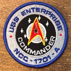 Puede incluir: Parche bordado con borde azul y el texto "USS Enterprise Commander NCC-1701-A". El parche presenta un centro negro con una insignia de Starfleet roja y amarilla y estrellas blancas.