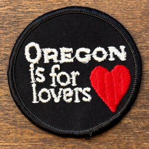 Puede incluir: Un parche redondo negro con las palabras "Oregon is for lovers" en escritura blanca. Un corazón rojo está en el lado derecho. El parche tiene un borde negro cosido.