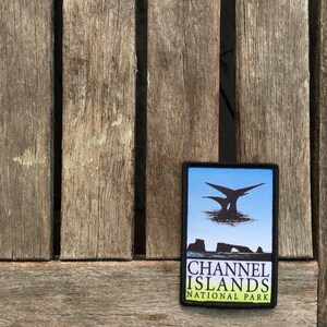 Op de afbeelding: Een zwart-witte patch met een blauwe achtergrond. De patch toont een silhouet van een eiland met een rotsboog en een vogel die erboven vliegt. De tekst "CHANNEL ISLANDS NATIONAL PARK" is gedrukt in witte letters op een groene achtergrond.
