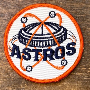 May include: Embroidered patch with the word "ASTROS" in navy blue, set against an orange and white design. The patch has an orange border and features a stadium and orbiting elements.