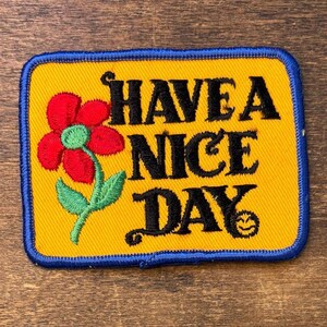 May include: A rectangular embroidered patch with a yellow background and a blue border. It features a red flower with a green center and stem on the left. The black text reads HAVE A NICE DAY, with a small smiley face icon at the end of the text.