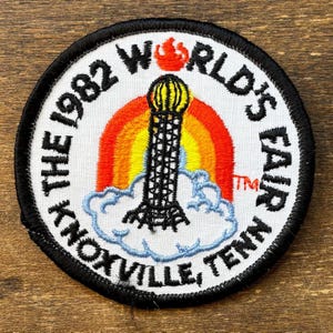 May include: Embroidered patch with a black border featuring text and graphics. The text reads "The 1982 World's Fair Knoxville, Tenn." A rainbow and a tower are in the center. The patch is circular and has a white background.