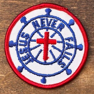 Può includere: Toppa ricamata circolare con una croce rossa e le parole "NEVER FAILS" in blu. La toppa ha uno sfondo bianco, un bordo rosso e un design a ruota di nave blu.