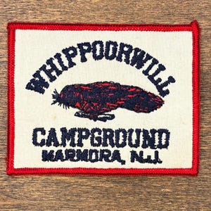 Może przedstawiać: Haftowana naszywka z czerwoną obwódką i kremowym tłem. Naszywka zawiera napis "WHIPPOORWILL CAMPGROUND MARMORA, N.J." w kolorze granatowym, wraz z haftowanym wizerunkiem ptaka w kolorze czerwonym i granatowym.