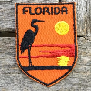 Puede incluir: Un parche bordado en forma de escudo con fondo naranja y borde negro. El texto FLORIDA está bordado en negro en la parte superior. Una garza negra se encuentra sobre una línea horizontal, con un sol amarillo y nubes rojas sobre un reflejo amarillo.