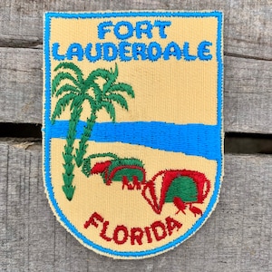 Puede incluir: Un parche bordado vintage que presenta una palmera, una masa de agua azul y dos tortugas marinas rojas y verdes. El parche es amarillo con ribete azul y dice "FORT LAUDERDALE FLORIDA".