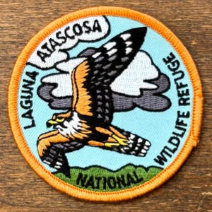 May include: Circular embroidered patch depicting a bird of prey in flight, with orange, black, and white plumage. The patch includes the text "Laguna Atascosa National Wildlife Refuge" and is set against a blue and green backdrop.
