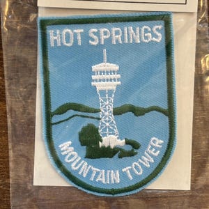 Könnte beinhalten: Ein blauer und grüner gestickter Aufnäher mit dem Text "HOT SPRINGS MOUNTAIN TOWER" und einer weißen Silhouette eines Turms.