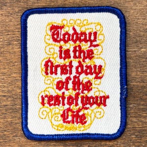 May include: Embroidered patch with a white background and a blue border. The patch features the text "Today is the first day of the rest of your life" in red and gold lettering. The design has a vintage aesthetic.