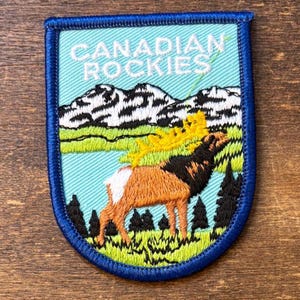 May include: Embroidered patch with a blue border featuring a mountain scene and an elk. The patch reads "CANADIAN ROCKIES" in white text. The elk is brown and black, with yellow antlers, standing in a green field with trees.
