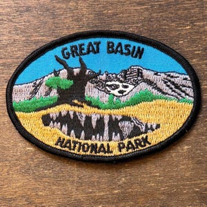May include: An oval embroidered patch featuring a blue sky, grey mountains, a dark brown tree, and green foliage above a yellow field. The text GREAT BASIN is embroidered in black at the top, and NATIONAL PARK is embroidered in black at the bottom.