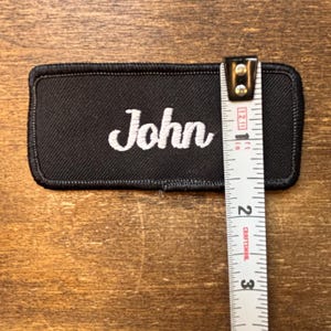 以下が含まれることがあります： 黒い長方形のパッチ。白い筆記体で「John」と刺繍されています。パッチの長さは約7.6cmで、定規で測定されています。