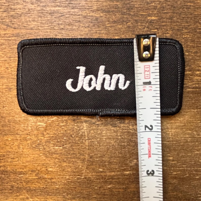 以下が含まれることがあります： 黒い長方形のパッチ。白い筆記体で「John」と刺繍されています。パッチの長さは約7.6cmで、定規で測定されています。