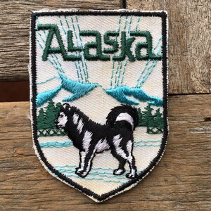 May include: White embroidered patch with a black and white husky dog standing in front of a mountain range. The text "Alaska" is embroidered in green above the mountains.