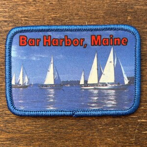 以下が含まれることがあります： 青い縁取りと赤い文字で「Bar Harbor, Maine」と書かれた長方形のパッチ。青い空の下、水面に浮かぶヨットの風景が描かれています。お土産やコレクションアイテムとして使用される可能性があります。