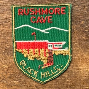 Könnte beinhalten: Ein grün-goldener, gestickter Aufnäher mit den roten Worten "RUSHMORE CAVE". Der Aufnäher zeigt ein rotes Gebäude, weiße Berge und die goldenen Worte "BLACK HILLS".