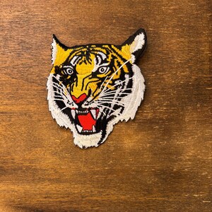 May include: An embroidered patch featuring the head of a tiger with yellow, black, and white fur. The tiger has a snarling expression with an open mouth showing white teeth and a bright red tongue, with black stripes across its face and ears.