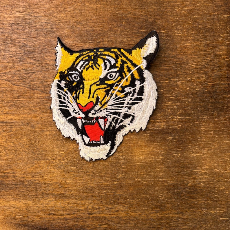 May include: An embroidered patch featuring the head of a tiger with yellow, black, and white fur. The tiger has a snarling expression with an open mouth showing white teeth and a bright red tongue, with black stripes across its face and ears.
