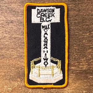 Op de afbeelding: Geborduurde patch met een wegwijzer met de tekst "DAWSON CREEK B.C." en "MILE 0 ALASKA HWY" op een zwarte achtergrond. De patch heeft een gele rand en een gedetailleerd ontwerp van de wegwijzer.