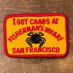 Op de afbeelding: Geborduurde patch met een gele achtergrond en een rode rand. De tekst "I GOT CRABS AT FISHERMAN'S WHARF SAN FRANCISCO" is rood. Een zwarte krabafbeelding staat in het midden.
