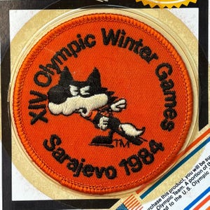 May include: Round orange patch with embroidered text and a cartoon fox mascot. The patch reads "XIV Olympic Winter Games Sarajevo 1984". The fox is black and white with an orange outfit. A vintage collectible.