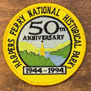 Op de afbeelding: Een cirkelvormige gele patch met geborduurde tekst en afbeeldingen. De patch leest "HARPERS FERRY NATIONAL HISTORICAL PARK" rond de rand. Het midden toont "50th ANNIVERSARY" en een landschap met een kerk en rivier. Data 1944-1994 staan onderaan.