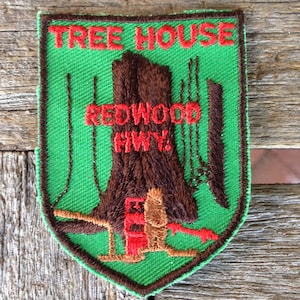 May include: A shield-shaped embroidered patch with a bright green background and a brown border. It features a large brown tree trunk, thin brown tree outlines, and red embroidered text that reads TREE HOUSE and REDWOOD HWY.