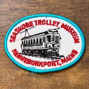 May include: An oval embroidered patch with a white background and a bright blue border. It features a black thread illustration of a trolley car and the red embroidered text "SEASHORE TROLLEY MUSEUM" and "KENNEBUNKPORT, MAINE" in a circular layout.