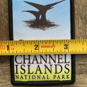 Op de afbeelding: Een zwart-witte patch met de tekst "Channel Islands National Park" en een afbeelding van een boom met drie takken. De patch wordt gemeten met een gele meetlint.