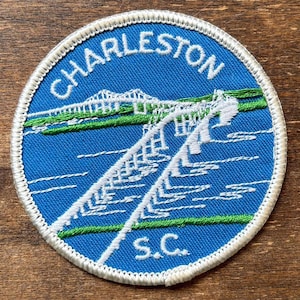 May include: Circular patch with a blue background featuring the word "CHARLESTON" in white, arched across the top. A white bridge design is in the center, with green accents. The text "S.C." is at the bottom, and the patch has a white border.