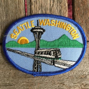 May include: Embroidered patch featuring the Seattle, Washington skyline. The design includes the Space Needle, a monorail, a sunset, and the text "Seattle Washington" in yellow on a blue background, with a blue border.