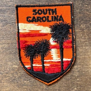 Op de afbeelding: Geborduurde patch met een zonsondergang, palmbomen en de tekst "SOUTH CAROLINA". De patch is oranje en zwart, schildvormig en waarschijnlijk voor een jas of tas. Het ontwerp roept een warm, tropisch gevoel op.