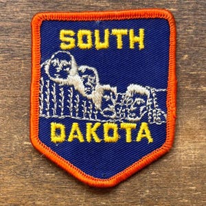 May include: A blue and orange embroidered patch featuring the words "SOUTH DAKOTA" in yellow. The patch depicts a white embroidered image of Mount Rushmore. The patch has an orange border.