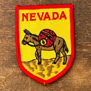 Könnte beinhalten: Gestickter Aufnäher mit dem Wort "NEVADA" in Rot über einem braunen Esel, der einen rot-schwarzen Rucksack trägt. Der Aufnäher hat einen gelben Hintergrund und einen roten Rand.
